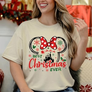 May include: A cream-colored short-sleeve t-shirt featuring a graphic of black mouse ears with a red and white polka dot bow. The design includes a black castle silhouette, a red Santa sleigh, green holly, and the text Best Christmas EVER.
