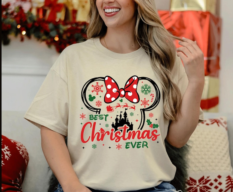 May include: A cream-colored short-sleeve t-shirt featuring a graphic of black mouse ears with a red and white polka dot bow. The design includes a black castle silhouette, a red Santa sleigh, green holly, and the text Best Christmas EVER.