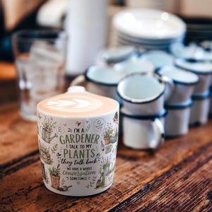 May include: A white ceramic mug filled with a latte featuring leaf-patterned foam art. The mug is decorated with illustrations of gardening tools and plants, and the text: I'M A GARDENER I TALK TO MY PLANTS they talk back WE HAVE A WHOLE CONVERSATION SOMETIMES.