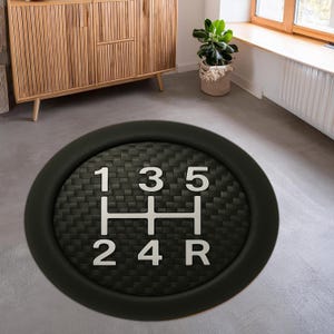 Könnte beinhalten: Runder, schwarzer Fußbodenbelag mit einem weißen Schaltschema in der Mitte. Das Schema zeigt die Zahlen 1, 2, 3, 4, 5 und R. Der Hintergrund des Musters ist ein schwarzes Karbonfasergewebe.