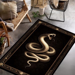 May include: A rectangular black rug with a detailed illustration of a golden snake with its mouth open. The snake is coiled on the rug, which has a decorative border with floral accents. The rug is placed on a concrete floor.