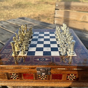 Puede incluir: Un juego de ajedrez decorativo con un tablero de ajedrez de madera con incrustaciones y piezas de ajedrez doradas y plateadas. El tablero presenta un patrón de cuadros blancos y negros. Las piezas de ajedrez están dispuestas en el tablero, listas para jugar. El juego está sobre una superficie de madera.