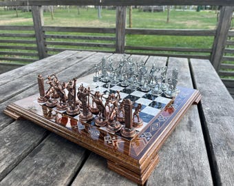 Op de afbeelding: Een decoratief schaakspel met een houten bord en gedetailleerde metalen schaakstukken. De stukken zijn zilver en brons, gerangschikt op een zwart-wit geblokt bord. Het bord heeft een decoratieve houten lijst.