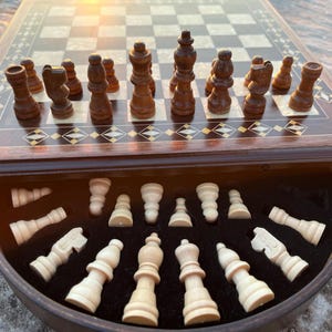 May include: A wooden chess set with a dark brown board and inlaid decorative border. The chess pieces are dark brown and light beige, with some pieces arranged on the board and others stored in a drawer below.