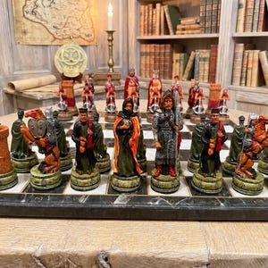 May include: A detailed chess set featuring hand-painted resin pieces depicting medieval figures, including knights on horseback, robed characters, and tall towers. The pieces are arranged on a rectangular board with a dark, marbled surface finish.