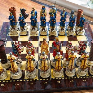 May include: A chess set featuring hand-painted resin pieces on a polished wooden board. One side features blue-clad figures and brown towers, while the other side displays cream-colored crusader-style figures with red crosses and brown towers.