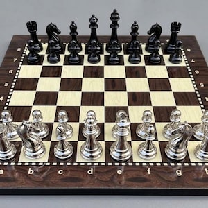 Op de afbeelding: Een schaakspel met een houten bord met afwisselend donkerbruine en lichtcrèmekleurige vakjes, gelabeld met de letters a tot en met h en de cijfers 1 tot en met 8. De set bevat een volledige set gepolijste zwarte en metallic zilveren schaakstukken.