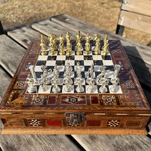May include: A decorative chess set with gold and silver pieces on a checkered board. The wooden box is inlaid with intricate patterns and a metal clasp. The chess pieces are arranged on the board, ready for a game.