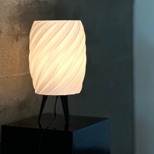 May include: A white ceramic table lamp with a unique, swirling design. The lamp has a black metal base with three legs and a cord.