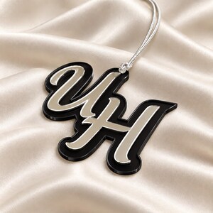 Può includere: Un ornamento in acrilico a strati che presenta le lettere UH in corsivo. Il design è composto da uno strato di base nero con uno strato superiore argentato riflettente, sospeso da un sottile cordino argentato fissato alla lettera H.
