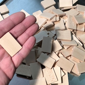 May include: A pile of unfinished, natural wood rectangular tiles. The tiles are approximately 1 inch by 2 inches. One tile is being held in a hand.