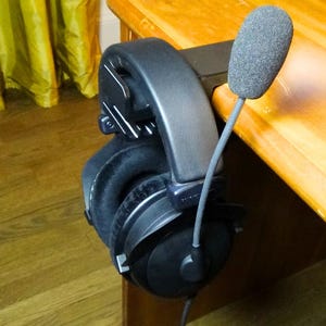 May include: A black headset with a microphone attached to a desk. The headset has a black headband and earcups, and the microphone is a small, black, round microphone.