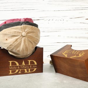 May include: Three baseball caps in shades of tan, gray, and maroon rest on a wooden box. The box is engraved with the word "DAD" and the names "Molly & Tiffany & Jon". A second wooden box with a deer antler design is also visible.