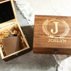 May include: A wooden box with a hinged lid, engraved with the letter "J" in a wreath and the name "JOSLYN". The box is open, revealing a brown leather flask inside.