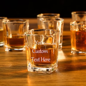 Op de afbeelding: Een set glazen shotglazen gevuld met amberkleurige vloeistof. Een shotglas heeft de tekst "Your Custom Text Here" gegraveerd aan de zijkant.