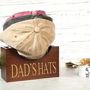 Puede incluir: Un perchero de madera para sombreros con el texto "DAD'S HATS" en la parte delantera. Tres gorras de béisbol están apiladas en la parte superior del perchero. Las gorras son beige, grises y rosas.