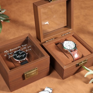 May include: Wooden watch display boxes with glass lids. One box contains a watch with a black band, and the other has a watch with a brown leather band. The boxes have a brown velvet interior.