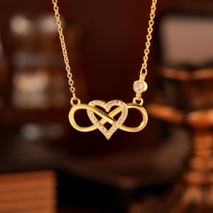 May include: A gold necklace with an infinity symbol and a heart, set with small, clear gemstones. The chain is fine, with a single gemstone near the clasp. The design is elegant and romantic, suitable for gifting. The necklace is a symbol of love.