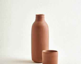 May include: A tall, cylindrical terracotta bottle with a narrow neck and a matching short, wide-mouthed terracotta cup. Both items feature a matte, earthy reddish-brown finish with a smooth, unglazed texture and simple, clean structural lines.