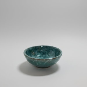 Puede incluir: Un pequeño cuenco redondo de cerámica con un esmalte moteado en tonos verde azulado, turquesa y azul oscuro. Las superficies interior y exterior muestran un patrón variado y salpicado con sutiles acentos marrones en el borde curvo.