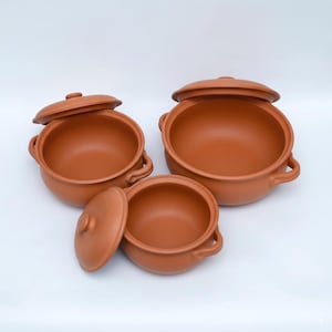 May include: A set of three terracotta cooking pots with matching lids. Each pot features a rounded body, two side handles, and a smooth, matte finish in a warm reddish-brown clay color. The lids have small central knobs for easy handling.