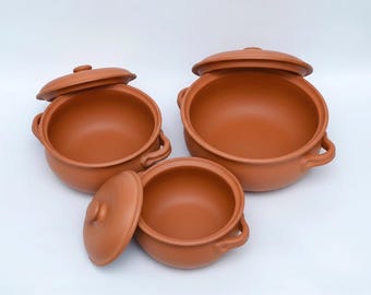 May include: A set of three terracotta cooking pots with matching lids. Each pot features a rounded body, two side handles, and a smooth, matte finish in a warm reddish-brown clay color. The lids have small central knobs for easy handling.