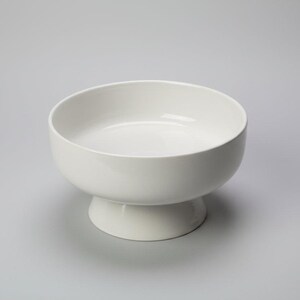 Pode incluir: Uma taça de cerâmica branca sólida com um acabamento liso e brilhante. A taça apresenta uma bacia circular larga que se estreita para uma base robusta tipo pedestal, criando uma silhueta equilibrada e elevada para esta peça de servir.