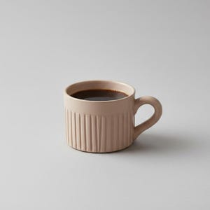 Op de afbeelding: Een lichtbeige keramische mok met een cilindrische vorm en een verticaal geribbelde textuur aan de buitenkant. De mok heeft een gebogen handvat aan de rechterkant en is gevuld met donkerbruine koffie, met een gladde, matte afwerking over het geheel.