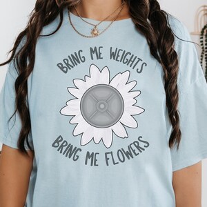 Puede incluir: Una camiseta azul claro con un gráfico de girasol blanco. El girasol tiene una placa de peso gris en el centro. El texto "Bring me weights bring me flowers" está impreso en la camiseta.