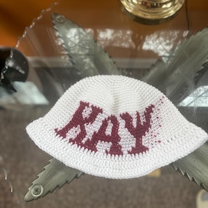 May include: White crocheted bucket hat with the name "Ray" embroidered in maroon.