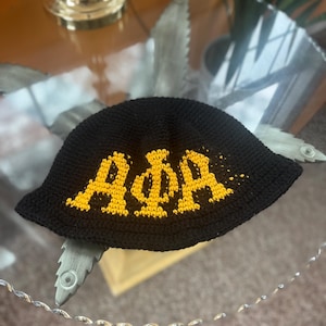 May include: Black crochet bucket hat with gold lettering spelling out "AΦA". The hat is displayed on a glass surface. The hat is a stylish accessory.