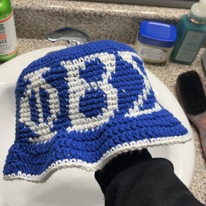 May include: A blue and white crochet bucket hat. The hat features a blue base with a white pattern and a white trim along the brim. The hat is handmade and appears to be made of yarn.