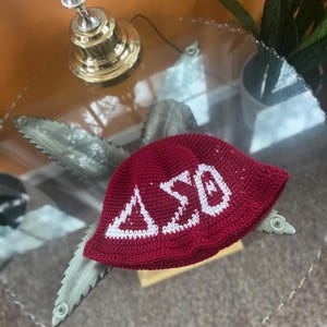 May include: A red crocheted hat with a white embroidered Delta Sigma Theta sorority logo.