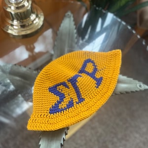 Könnte beinhalten: Ein handgefertigter, gelber Häkelhut mit den Buchstaben "ΣΙΡ" in Blau. Der Hut ist aus einem strukturierten Garn gefertigt und hat eine abgerundete Form. Der Hut liegt auf einer Glasoberfläche.
