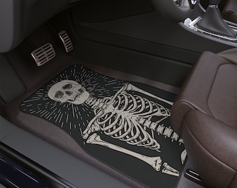 May include: Black car floor mat with a white skeleton graphic. The skeleton is sitting with its head tilted back and its arms outstretched. The graphic is surrounded by a black circle with white rays emanating from it.