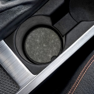 Puede incluir: Un portavasos negro para coche con un posavasos redondo con un diseño gris en forma de telaraña. El posavasos está dentro del portavasos, rodeado de plástico negro y un borde plateado. El interior del coche es visible.