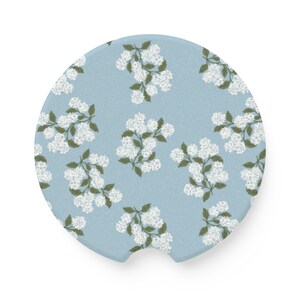 May include: A round blue car coaster with a white floral pattern. The coaster has a cutout in the bottom for easy gripping.