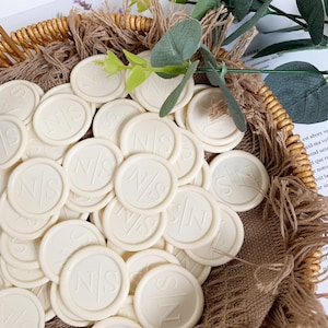 Könnte beinhalten: Ein Korb gefüllt mit weißen runden Wachssiegeln mit den eingeprägten Initialen "NS".