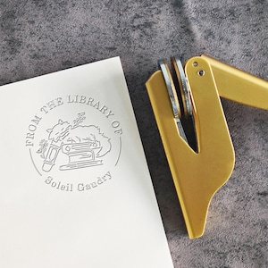 May include: A gold-colored self-inking stamp with a circular design. The design features a book, a quill pen, and a cat. The text reads "From the Library of Soleil Gaudry".