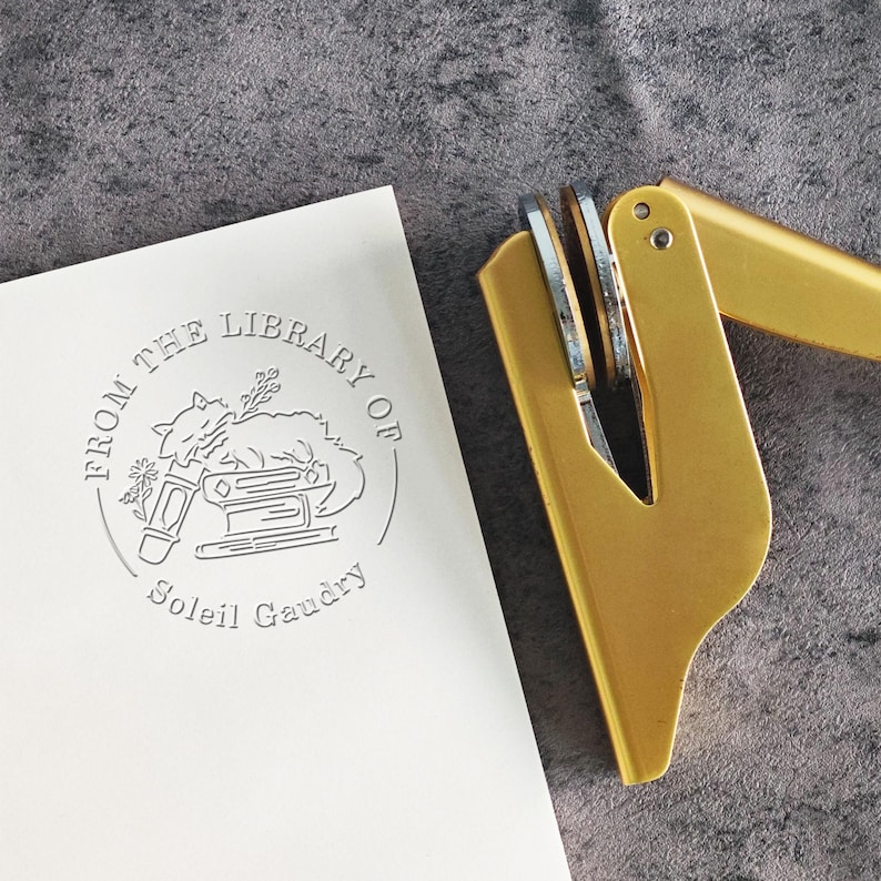 May include: A gold-colored self-inking stamp with a circular design. The design features a book, a quill pen, and a cat. The text reads "From the Library of Soleil Gaudry".
