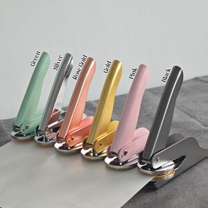 May include: A set of six metal stampers in different colors: green, silver, rose gold, gold, pink, and black. Each stamper has a handle and a round base with a raised design.