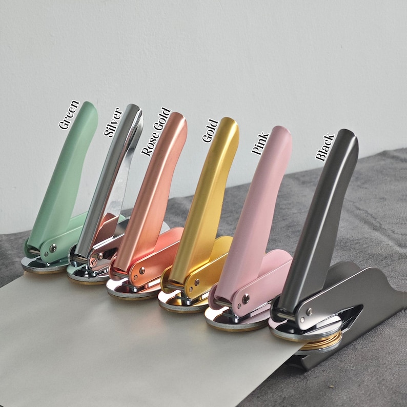 May include: A set of six metal stampers in different colors: green, silver, rose gold, gold, pink, and black. Each stamper has a handle and a round base with a raised design.