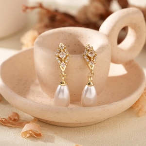 Pode incluir: Um par de brincos pendurados dourados com uma parte superior em forma de diamante adornada com cristais transparentes e um pendente de pérola branca em forma de pêra. Os brincos são exibidos em um pires de cerâmica bege.