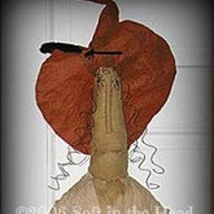 Puede incluir: Una muñeca de trapo hecha a mano vestida con un vestido negro y un gran sombrero naranja con una pluma negra. La muñeca sostiene una calabaza roja y negra.