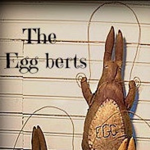 以下が含まれることがあります： 長い細い腕と脚を持つ、茶色のフェルト製の卵型のフィギュア2体。フィギュアは、グリッドパターンが描かれた白い背景に付けられています。画像の上部には黒い文字で「The Eggberts」と書かれています。