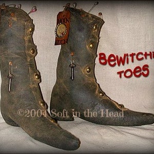 Puede incluir: Un par de botas grises oscuras de estilo vintage con puntas puntiagudas. Las botas presentan tachuelas de color latón, etiquetas decorativas y pequeños dijes de llave. El texto "Bewitchin' toes" está escrito en rojo. Las botas parecen hechas a mano.