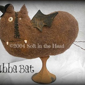 Op de afbeelding: Een bruine vleermuis van vilt met een zwarte vilthoed en vleugels, staand op een houten voet. De vleermuis heeft een knoopoog en een gestikte mond met witte tanden. De tekst "Bubba Bat" staat onder de vleermuis. ©2004 Soft in the Head