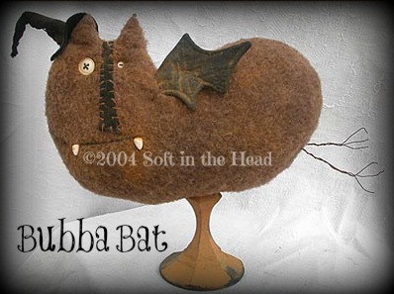 May include: A brown felt bat with a black felt hat and wings, standing on a wooden base. The bat has a button eye and a stitched mouth with white teeth. The text "Bubba Bat" is written below the bat. &copy;2004 Soft in the Head