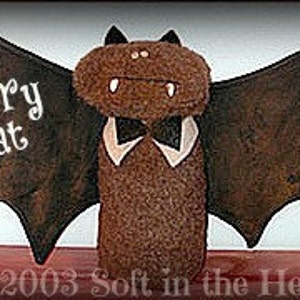 Op de afbeelding: Een bruine vleermuispop van vilt met grote zwarte vleugels, een witte kraag en een zwarte strik. De pop heeft een gestikt gezicht met twee zwarte ogen en een grijns. De tekst "Belfry Bat" is in wit op de vleugels gestikt.