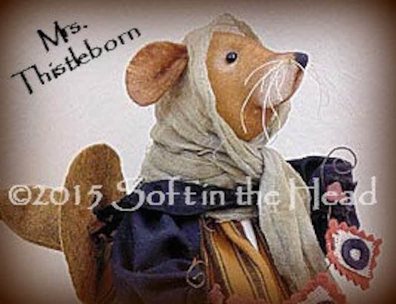 Peut inclure: Poup&eacute;e souris en tissu faite &agrave; la main nomm&eacute;e "Mrs. Thistleborn", portant un foulard et une robe, et tenant un petit sac. La poup&eacute;e a un corps beige, des oreilles marron et de longues moustaches blanches. Le texte "Mrs. Thistleborn" et "&copy;2015 Soft in the Head" sont visibles.